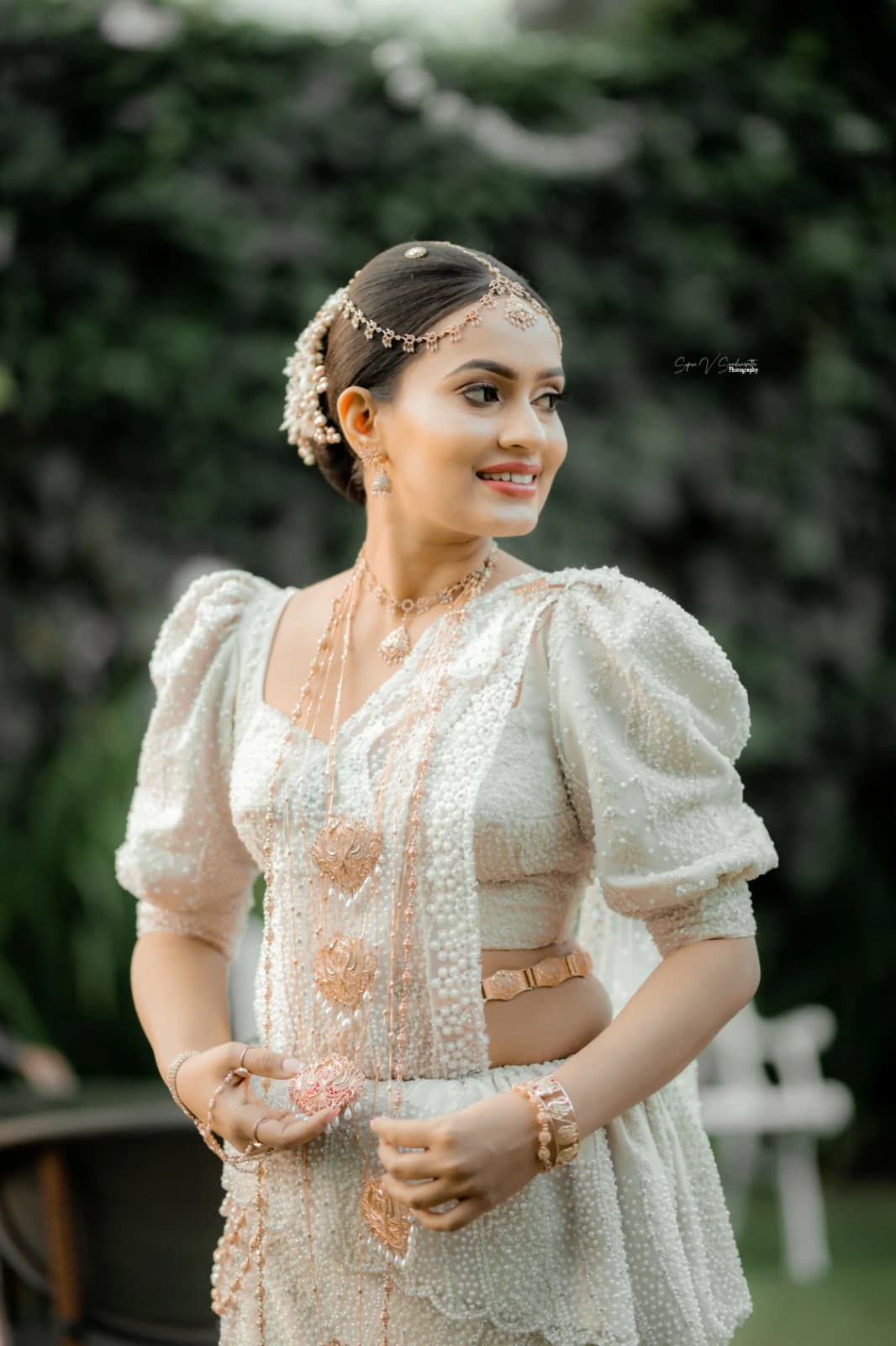 Bride in an embroidered ivory gown and gold headpiece, smiling in a leafy garden